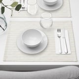 Mesa con 4 alfombras GOKVÄLLÅ de cor crema, cada unha con pratos brancos, vasos e pratería. As alfombras son menos anchas que os pratos que conteñen.