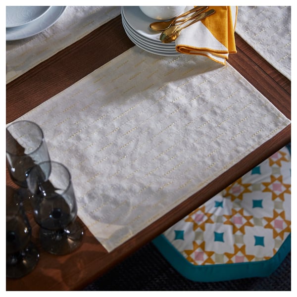Placemat rectangular amarelo dourado sobre mesa de madeira, con pratos brancos, cubertas de ouro e vasos transparentes.