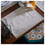 Placemat rectangular amarelo dourado sobre mesa de madeira, con pratos brancos, cubertas de ouro e vasos transparentes.
