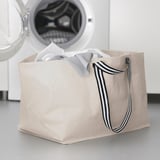 Bolsa de lavandaría beige GÖRSNYGG con mangos de raias, verticalmente xunto á lavadora, chea de roupa lixeira.