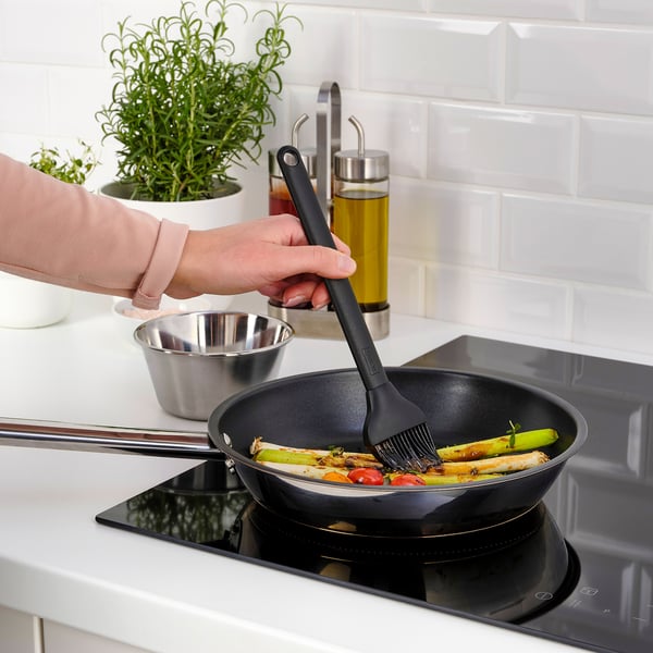 Persoa á grella de verduras usando cepillo GRILLTIDER en estufa negra, con herbas e aceite ao fondo.