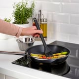 Persoa á grella de verduras usando cepillo GRILLTIDER en estufa negra, con herbas e aceite ao fondo.