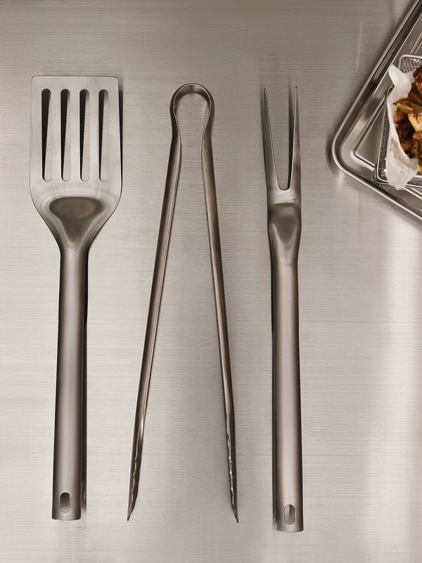 Tres ferramentas de grella de aceiro inoxidable: pinzas, espátula e garfo, colocadas sobre unha superficie metálica. Deseño elegante, práctico para grella.