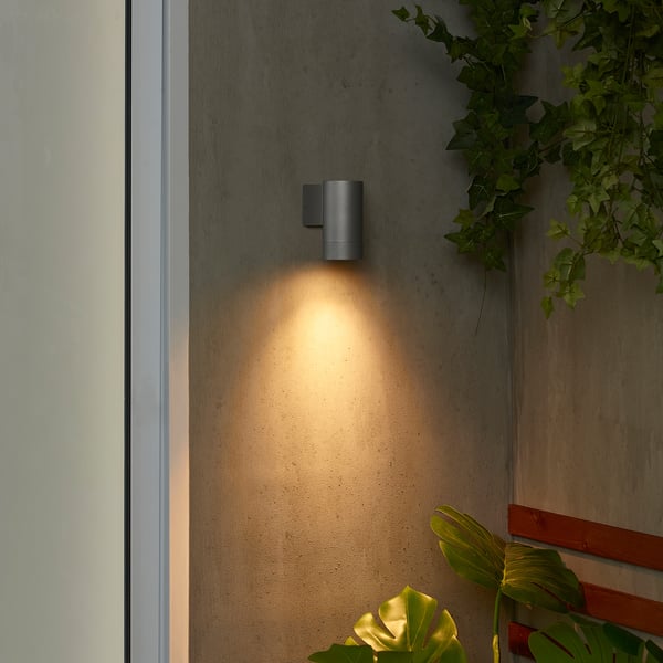 A pequena lámpada de parede GRÖNSPRÖT negra emite unha luz cálida xunto á porta branca, rodeada de plantas verdes, destacando o seu tamaño compacto e o seu deseño elegante.