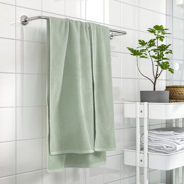 Baño moderno con gran toalla verde GULVIAL colgada, rodeado de baldosas brancas e unha pequena planta.