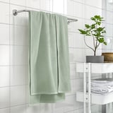 Baño moderno con gran toalla verde GULVIAL colgada, rodeado de baldosas brancas e unha pequena planta.