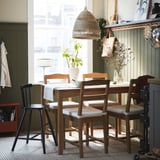 Acolledora zona de comedor con conxunto HÄGERNÄS de madeira, gran luz colgante, plantas e estantes decorados.