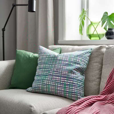Almofada decorativa HAMNKRASSING con raias no sofá, máis pequena, lámpada verde, planta detrás.
