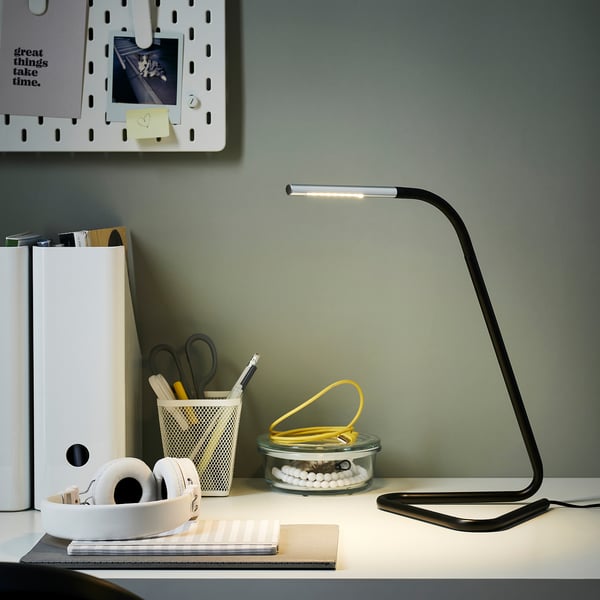 Configuración de escritorio moderna con lámpada HÅRTE negra, delgada e axustable. Auriculares brancos, papelería e notas ao lado. Fondo de parede verde.