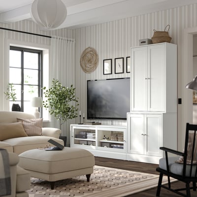 Sala de estar moderna con unidades de almacenamento HAVSTA branco, sofá beige, alfombra con patróns e papel de pantalla a raias; Artigos de televisión e decoración exhibidos.