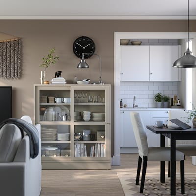 Interior moderno con estante de madeira HAVSTA en beige, exhibindo pratos e libros. Reloxo negro, cociña branca e comedor gris.