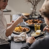 A persoa cociña pasta con salsa nunha tixola sobre unha estufa. Pratos e botellas sobre a mesa.
