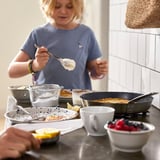 O neno cociña panqueiques nunha tixola antiadherente, rodeada de ingredientes e ferramentas.