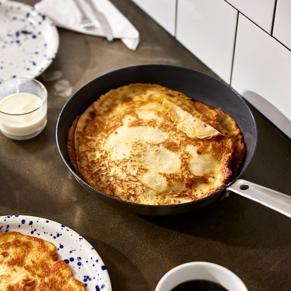 Unha tixola antiadherente con panqueica dourada aséntase nunha mesa, emparellada con pratos e crema.