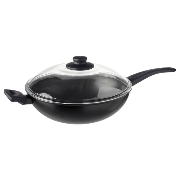 Un wok HEMLAGAD negro cunha tapa de vidro e dous mangos robustos. A súa superficie antiadherente e o seu deseño profundo convérteno ideal para cociñar sen derrames.