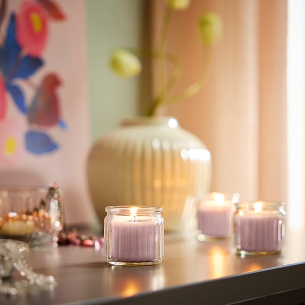 Pequenas velas rosas en recipientes de vidro sobre unha mesa, rodeadas de obxectos decorativos como un xarrón con flores e un paxaro pintado.