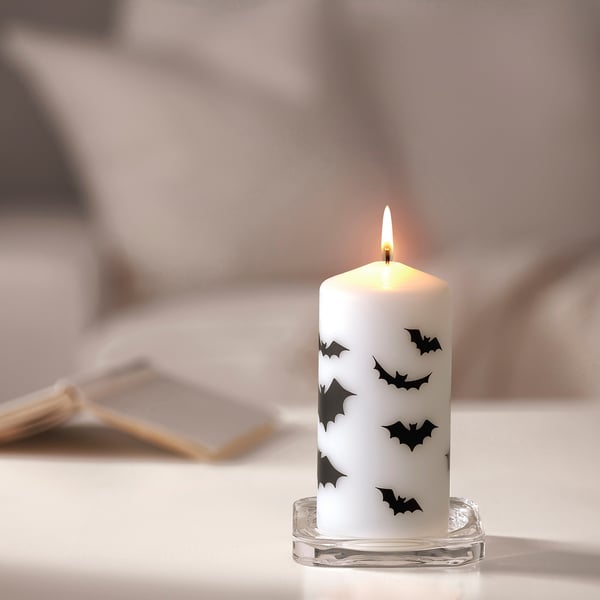A vela branca de Halloween HÖSTAGILLE, decorada con morcegos negros, está nun soporte de vidro transparente. É pequeno, colocado xunto a mistos nunha mesa.