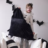 Neno con traxe de morcego de Halloween sostendo un coxín suave negro, sentado nun sofá branco con almofadas de raias brancas e negras.