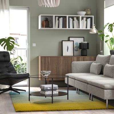 Sala de estar moderna con sofá gris, alfombra colorida, mesa de vidro, armario de madeira, cadeira de coiro, estantes con libros e paredes verdes claras.