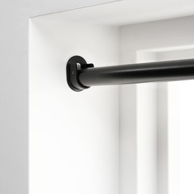 Soporte de barra de cortina negra montado en parede branca, parte do sistema HUGAD para colgar cortinas entre dúas paredes opostas.