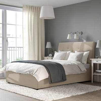 A cama beige IDANÄS cunha cabeceira inclinada, cortinas brancas e lámpadas de parede. A cama é a peza central da habitación, máis grande que outros elementos.