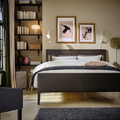 Dormitorio moderno con cama de madeira escura IDANÄS, roupa de cama branca, arte negra, estantes con libros e plantas.