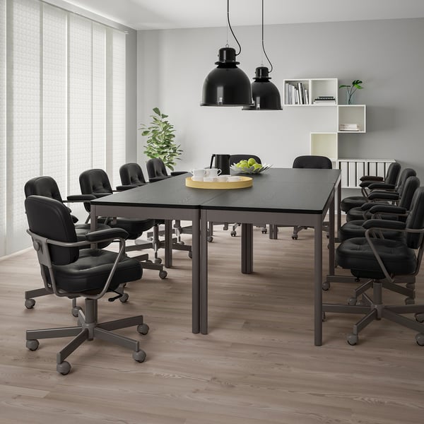 Sala de conferencias moderna con mesa IDÅSEN negra, cadeiras xiratorias, lámpadas colgantes, plantas e gran xanela para a luz natural.