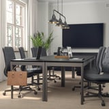 Sala de reunións moderna con mesa rectangular, cadeiras grises, luces colgantes, planta en pota e bolsa de coiro. Grandes fiestras.
