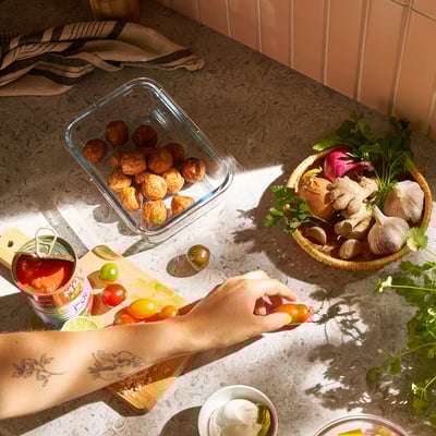 Unha persoa corta os tomates nunha táboa de corte xunto a verduras asadas e produtos frescos, mostrando a preparación e almacenamento de alimentos.