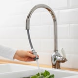 A persoa acende a billa da cociña cromada, a auga verte no lavabo con verduras de folla verde.