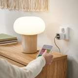 Unha persoa sostén un teléfono intelixente cunha aplicación de enchufe intelixente, controlando unha lámpada beige nunha mesa de madeira.