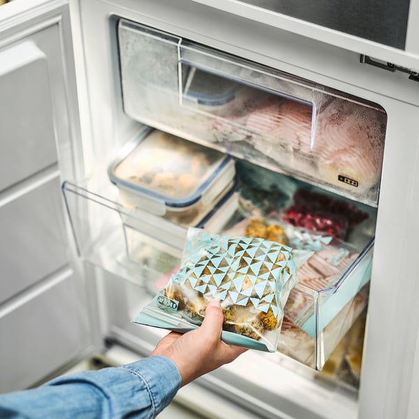 A man sostén unha bolsa de alimentos reutilizable etiquetada ISTAD que mostra o seu contido, colocada nun caixón de conxelador.