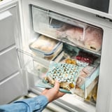 A man sostén unha bolsa de alimentos reutilizable etiquetada ISTAD que mostra o seu contido, colocada nun caixón de conxelador.