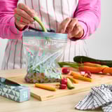 Persoa selando unha bolsa de verduras reutilizable con patrojo colorido nunha mesa de madeira rodeada de produtos frescos.