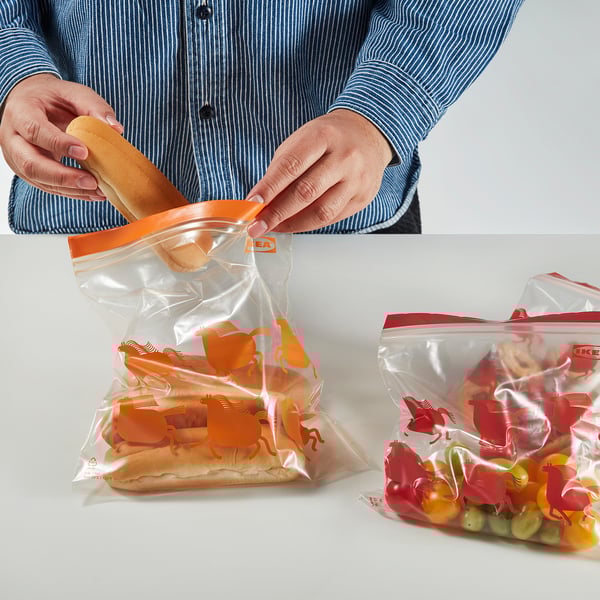 A persoa abre unha bolsa de plástico que contén un hot dog. Son visibles dúas bolsas, unha cun hot dog e outra con uvas.