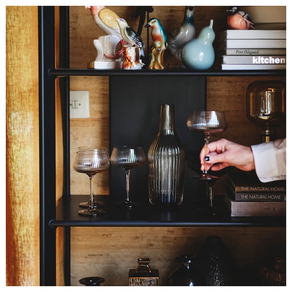 Estante con marco metálico negro e estantes de bambú. Decorado con vasos e figuriñas. Unha man que sostén un vaso de viño. Libros colocados no estante superior.