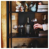 Estante con marco metálico negro e estantes de bambú. Decorado con vasos e figuriñas. Unha man que sostén un vaso de viño. Libros colocados no estante superior.