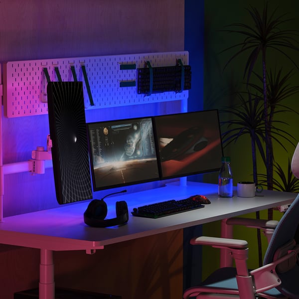 Escritorio moderno con monitores dobres, teclado, rato, auriculares para xogos e luces led. Cadeira de oficina rosa, planta de araña, espazo de traballo elegante.