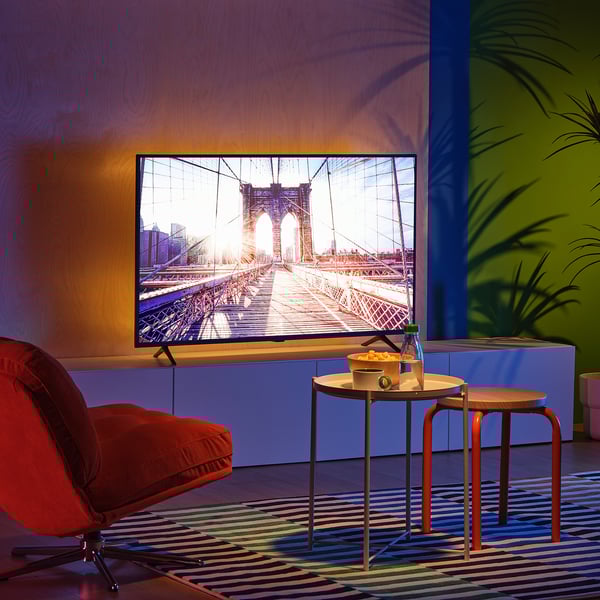 Sala de estar moderna con televisión de pantalla plana que mostra unha ponte. Cadeira vermella brillante, pequenas mesas redondas e alfombra de cores con plantas.