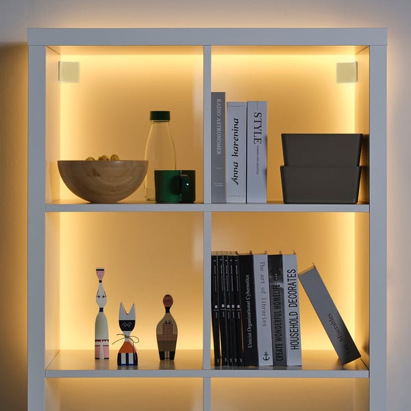 Unha estantería iluminada con libros, decoración e tiras de luz KABBLEKA . Os libros póñense en pé, a decoración varía en tamaño, a iluminación destaca os obxectos.