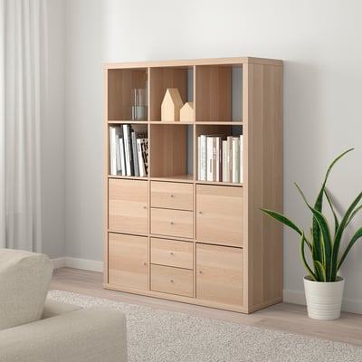 Unidade de estante KALLAX con libros, decoración e planta. Sofá beige parcialmente visible.