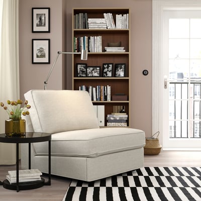 Espazo de estar acolledor con sofá de cama gris KIVIK de 1 asento, alfombra a raias, paredes rosas calmantes, estante de libraría e mesa lateral negra con vaso amarelo.