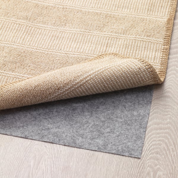 Alfombra tecida beige con patrojo texturizado, parcialmente dobrada sobre almofada de agarre gris.