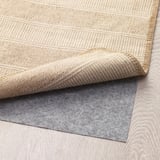 Alfombra tecida beige con patrojo texturizado, parcialmente dobrada sobre almofada de agarre gris.