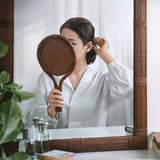 Muller que usa espello de ratán para maquillaxe. Alternativa con número de personaxes: muller con espello de maquillaxe de ratán.