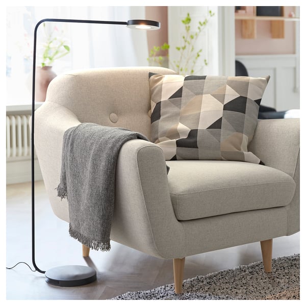 Elegante cadeira de brazos beige con lámpada moderna, almofada xeométrica, manta gris sobre tecido acolledor, patas de madeira, preto da fiestra con plantas.