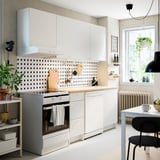 Pequena cociña branca con armarios KNOXHULT, polka dot backsplash, forno inoxidable, mesa de madeira, disposición eficiente, estantes de parede e zona de comedor.