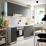 Cociña moderna con armarios grises, mostradores brancos e backsplash de puntos. As cadeiras negras da fiestra, as plantas e a decoración engaden calor.