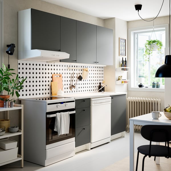 Cociña moderna con armarios grises KNOXHULT, tapa de traballo de mármore branco, electrodomésticos inoxidables, backsplash de puntos, plantas colgantes e cadeiras negras.