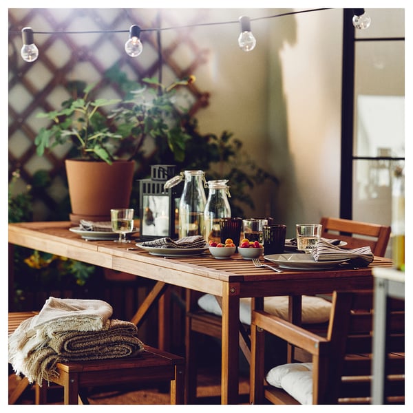 Conxunto de mesa ao aire libre con cadeiras de madeira, frascos KORKEN, vasos, pratos, cubertas, cubertas de froita e luces de corda.